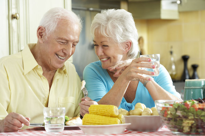 older dental patients smiling at each other while eating corn because they went to a periodontist for their implant supported dentures procedure.