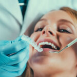 A dentist, wearing blue gloves, is holding a clear dental aligner and is about to place or adjust it in the mouth of a female patient, who appears relaxed and has her mouth open.